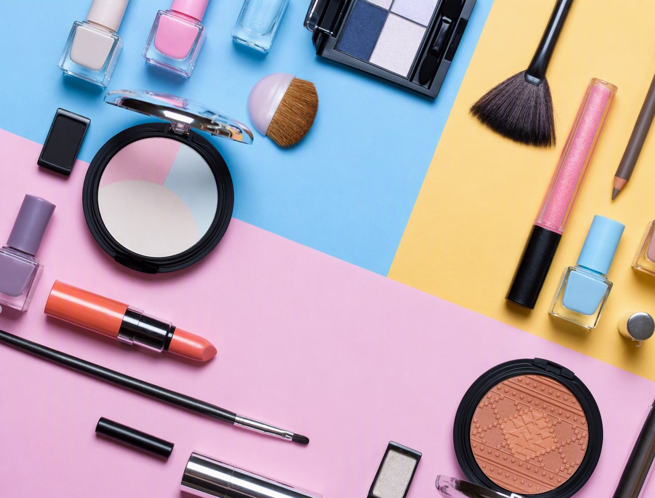 Colección completa de maquillaje sobre fondo tricolor: paletas, labiales, máscara, brochas y esmaltes